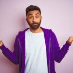 Young indian man wearing purple sweatshirt standing over isolated pink background smiling showing both hands open palms, presenting and advertising comparison and balance